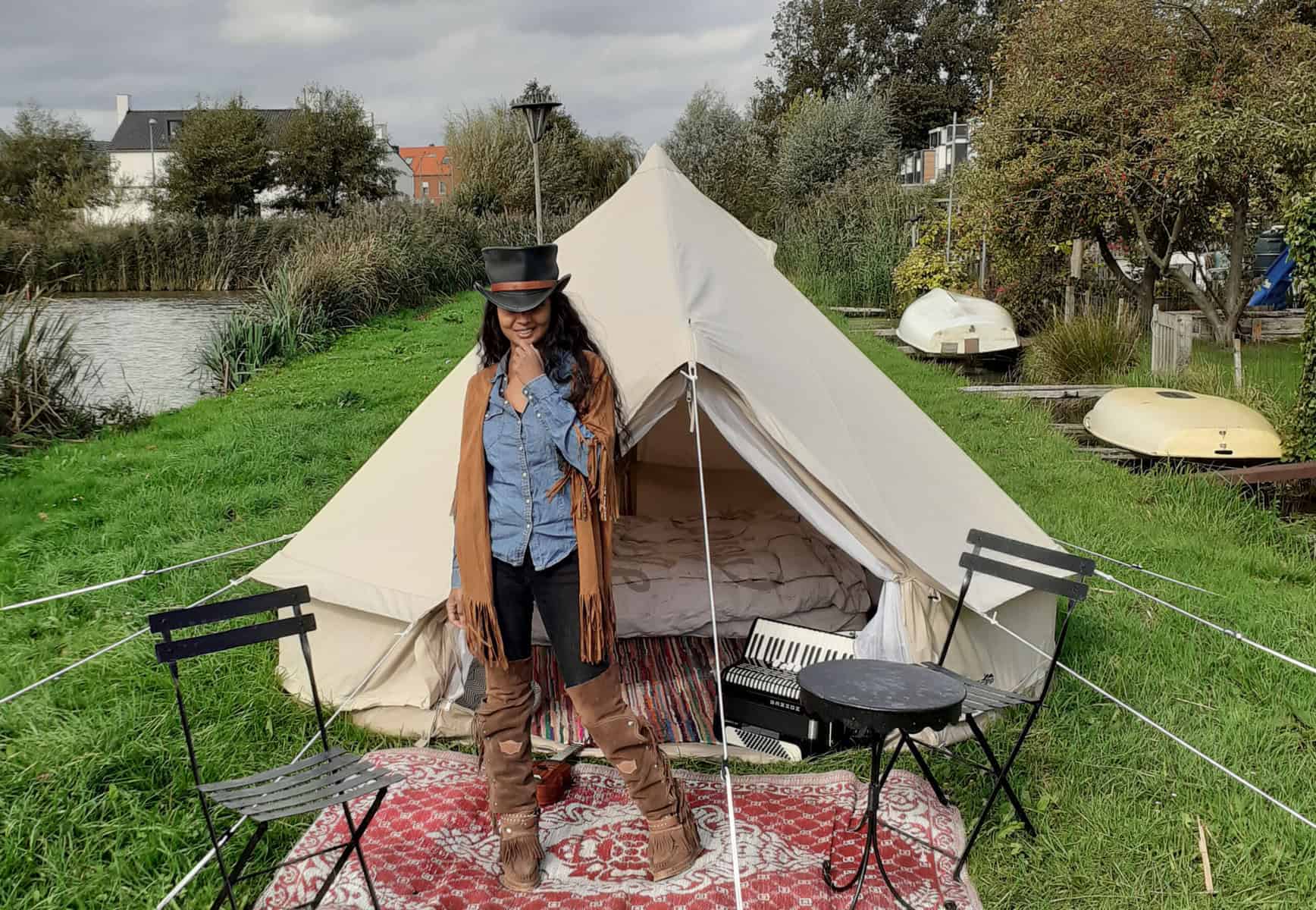 Stylische Frau im Cowboy-Outfit vor einem weißen Tipi-Zelt, auf einer Wiese neben einem Fluss in ruhiger, naturnaher Umgebung, mit Boote im Hintergrund, bei schönem, leicht bewölktem Wetter.