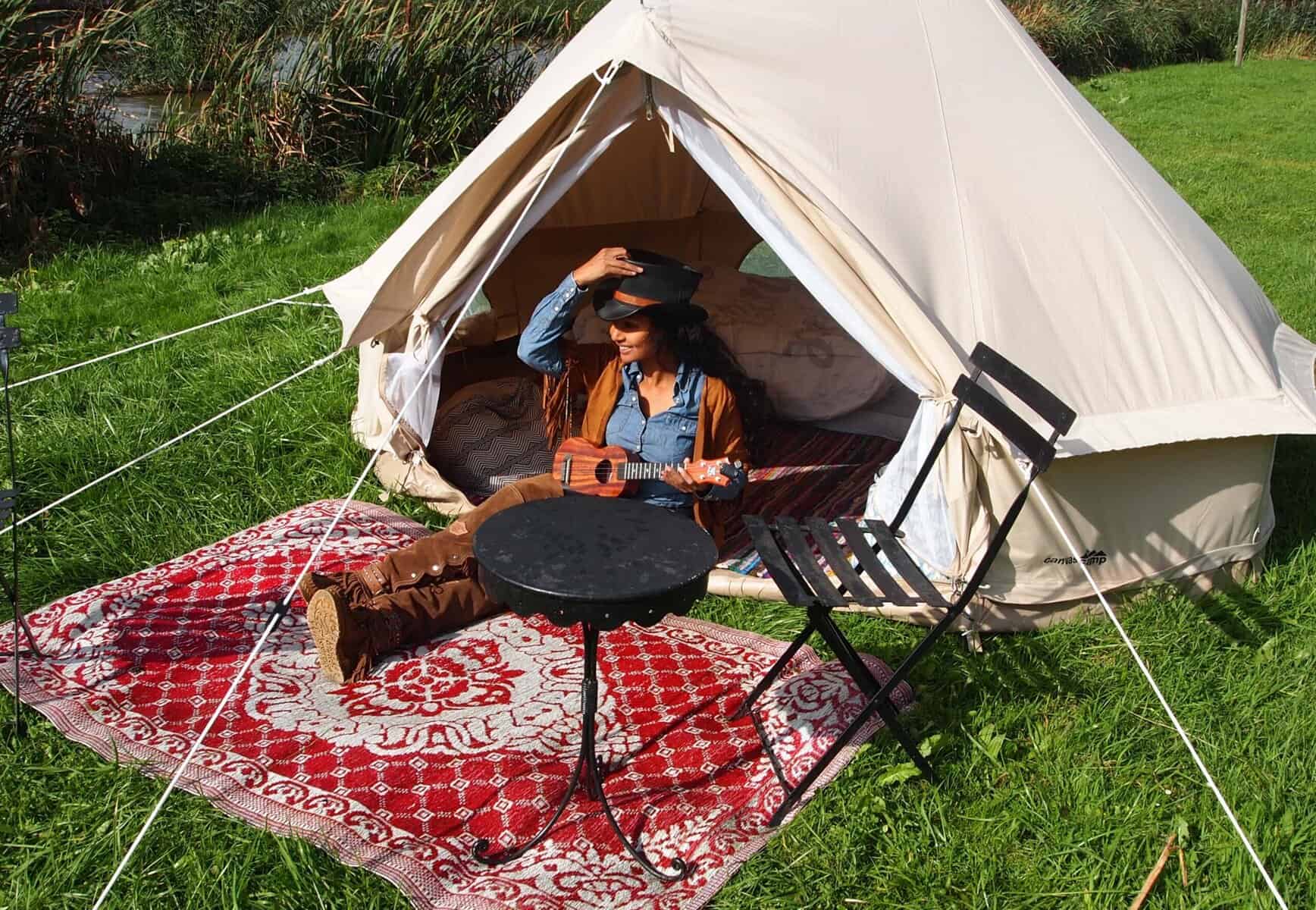 Gemütliche Zeltszene mit Frau, die Gitarre spielt, auf buntem Teppich im Grünen, entspannte Campingatmosphäre, Natur, Freizeit, Outdoor, Abenteuer, richtiges Campingzubehör, entspannter Tag am Wasser.