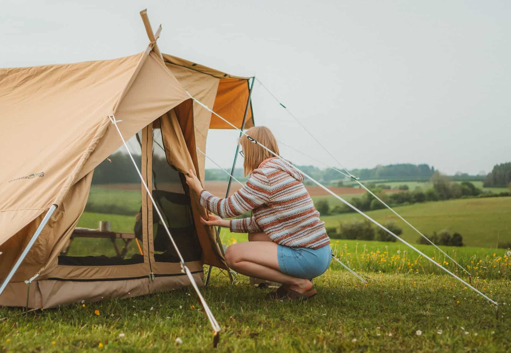 Vorderansicht einer Frau, die vor einem Campingzelt im Grünen kniet, das Zelt ist beige-braun, im Hintergrund weite grüne Wiesen und ein bewölkter Himmel, Szene beim Campingurlaub in der Natur.