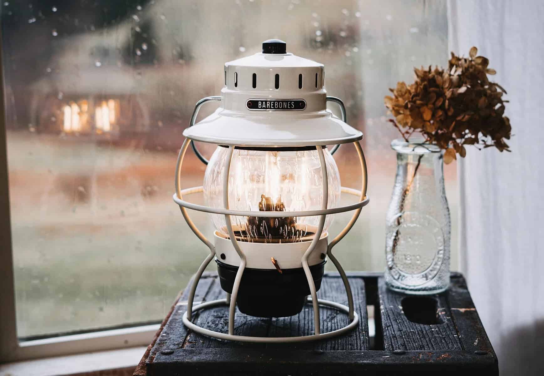 Leuchtende Petromax Laterne im Vintage Design auf Holztisch mit getrockneten Blumen in Vase, gemütliche Atmosphäre, ideal für Camping, Outdoor und stilvolle Dekoration.