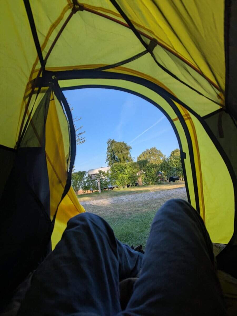 Hauptbild: Blick aus einem gelben Zelt auf eine sonnige Campingwiese mit Bäumen und Wolken.