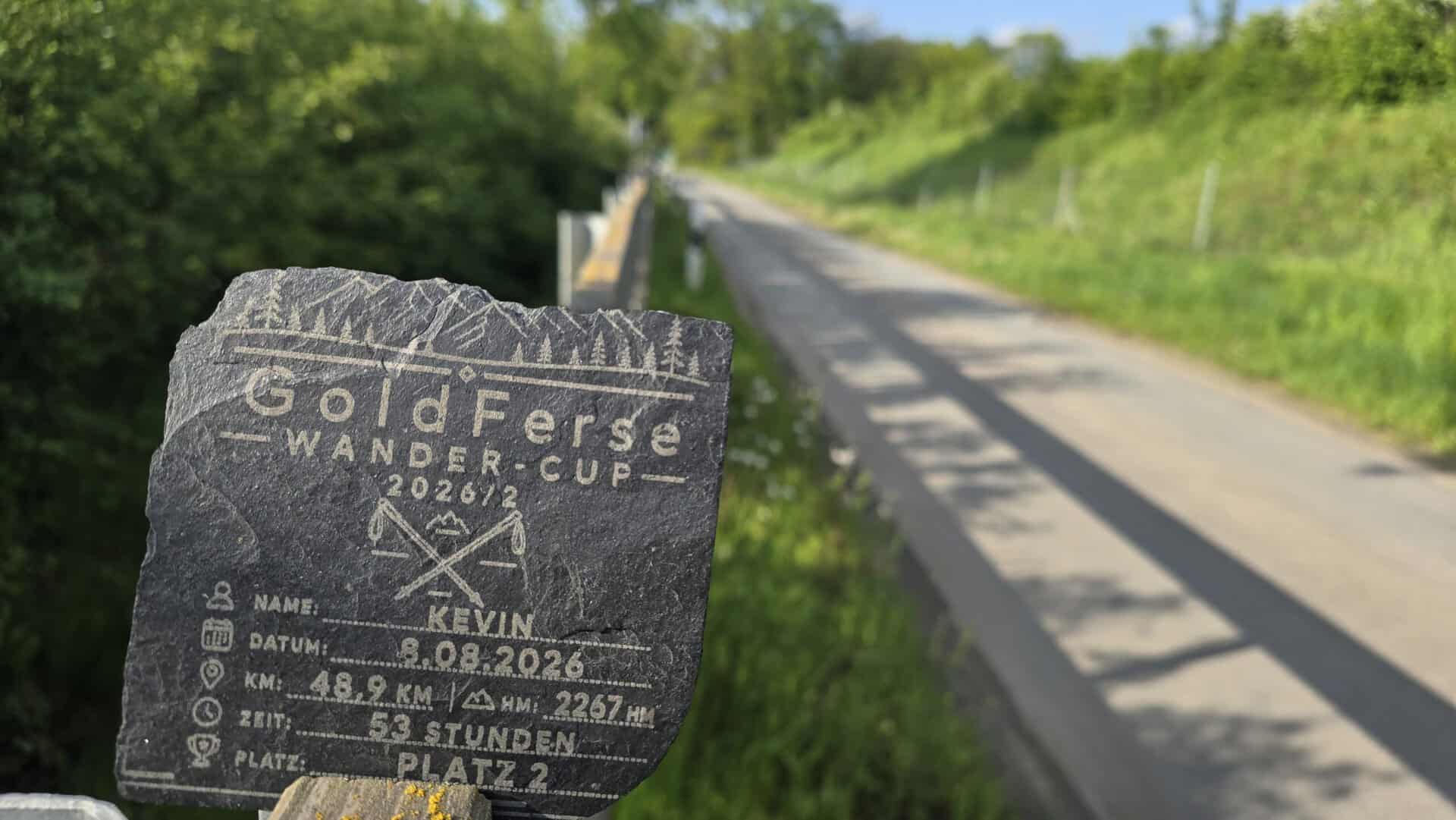 Wanderer mit graviertem GoldFerse Wander-Cup 2026 auf einem Stein, im Hintergrund eine ländliche Straße und grüne Natur, Symbol für Wandertouren und Naturerlebnisse.