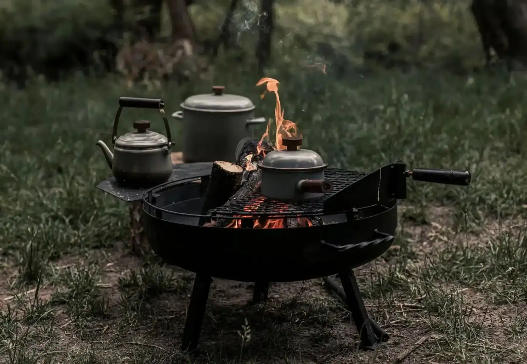 Kamin-Feuerstelle mit drei Teekannen auf einem Grill im Freien, ideal für Camping und Outdoor-Kochen, robustes Design für langlebigen Gebrauch.