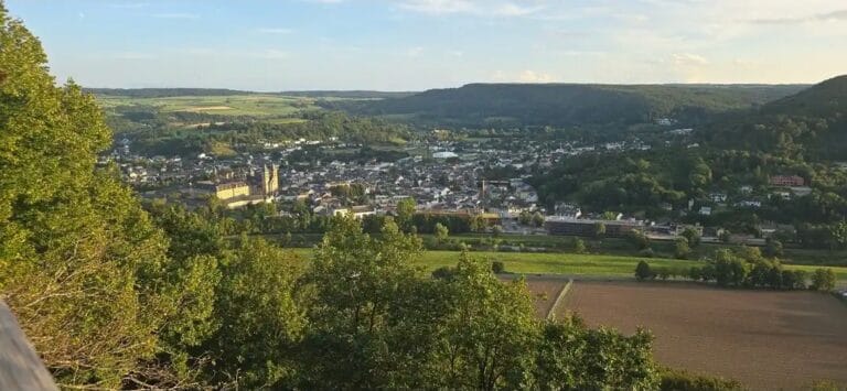 Weitblick über eine grüne Landschaft mit Fluss und Stadt im Hintergrund, umgeben von Hügeln, bei sonnigem Wetter.